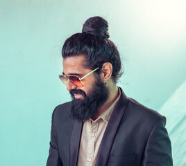 stylish man with a topknot hairstyle and a full beard is seen wearing a dark blazer and a light beige shirt underneath. He's smiling while looking down slightly off to his side