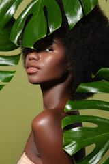 Peeking through lush tropical leaves, a young African American woman with radiant skin and curly afro hair poses gracefully on an olive background. Fashion beauty skincare portrait
