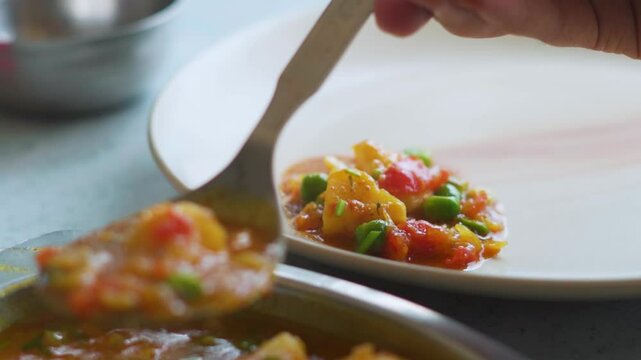 Closeup shot of man placing cooked Aloo mutter on a plate with the help of spoon for his lunch. Indian food background with copy space. Traditional Indian sabji or cooked Vegetables on a plate.