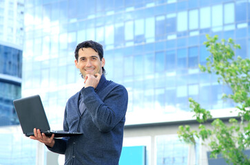 Fototapeta premium Business man stands in front of an office building with a laptop in hand.