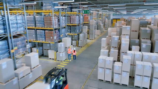 A bird's-eye view of a bustling warehouse reveals workers in orange vests navigating aisles filled with crates and boxes, highlighting the complex logistics of global supply chains.