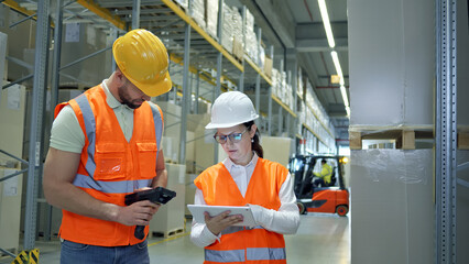 In a warehouse, two workers in orange vests and hard hats collaborate, using a tablet and a scanner, symbolizing efficient global supply chain logistics.