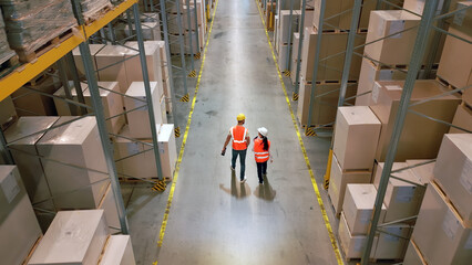 Aerial view of two workers in a vast warehouse, navigating between towering shelves of packages, symbolizing the complexity and scale of global logistics and supply chain management.