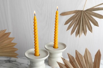 Burning beeswax candles in candlesticks on white marble table and dry palm leaves, closeup