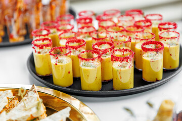 Colorful appetizer shot glasses filled with creamy yellow soup and garnished with herbs and red seasoning. Served on a black tray at a catered event, showcasing elegant presentation and attention to d