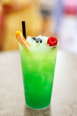 A vibrant green beverage served in a clear plastic cup with ice, garnished with a fresh orange slice, raspberry, and blueberries.