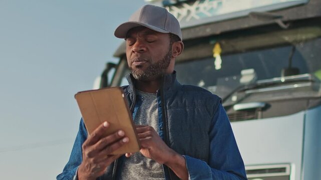 Focused African American male in gray cap interacting with tablet near front of truck. Scrolling or tapping screen while checking cargo information or managing digital delivery logs.