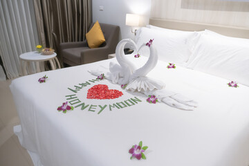 The honeymoon. Swans in the shape of hearts made of towels and a heart made of red rose petals on a snow-white bed in a hotel room as a symbol of love. Happy Honeymoon inscription made of green stems