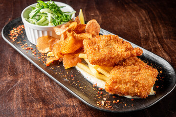 A plate featuring crispy fried fish fillets alongside golden French fries and homemade potato chips. A small dish of refreshing cucumber salad adds a vibrant touch to this hearty meal.