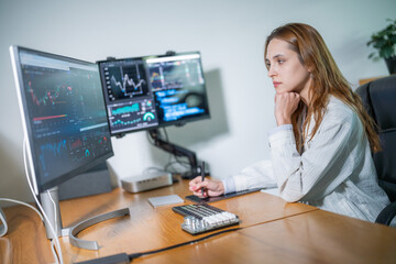 Woman analyzes market trends at a dual-monitor workstation during the day in a modern office...
