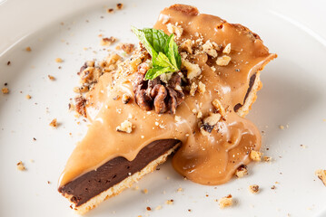 A slice of rich chocolate pie topped with caramel sauce, chopped nuts, and a fresh mint leaf. The dessert is beautifully presented, offering a decadent and inviting appeal.