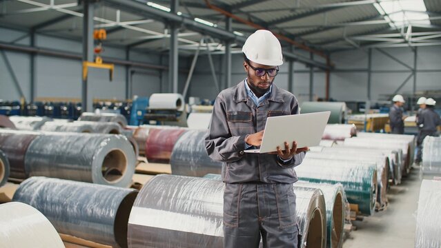 Serious African American engineer using laptop at workplace. Worker wearing helmet and uniform standing in large storage. Inspector of manufacture working with technology device at warehouse.