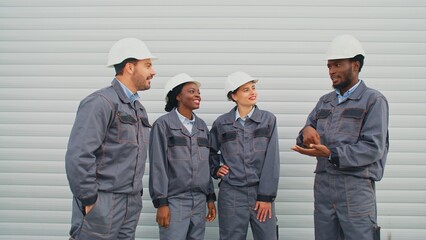 Zooming out from multiethnic workers communicating near large factory. Team of engineers positively communicating during working time. People with uniforms and helmets adjusting their helmets outside.