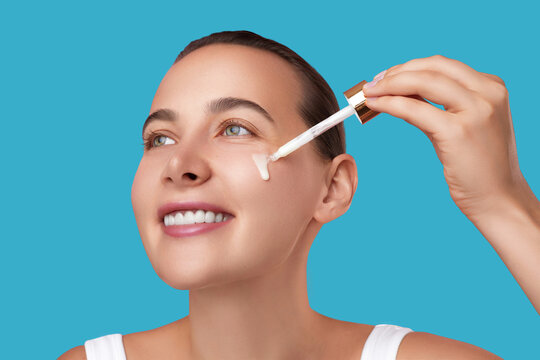 Applying serum with a dropper, a young woman with glowing skin smiles and looks up, posing against a bright blue background. Skincare, hydration and rejuvenation concept.