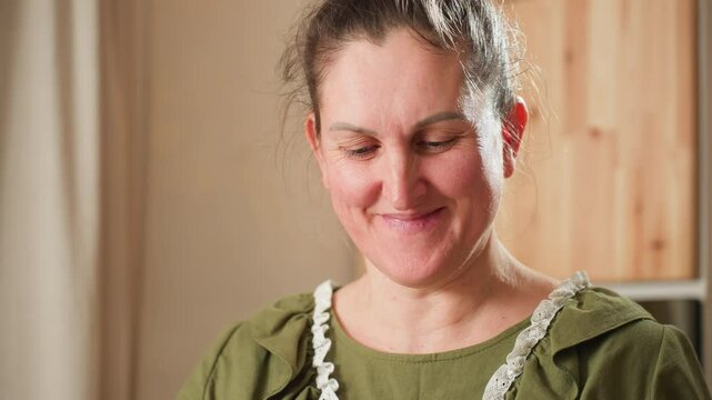 White woman in green ruffle dress and white apron smiling warmly while dancing joyfully in cozy kitchen, holding mixing bowl in hand near stand mixer, radiating lighthearted happiness