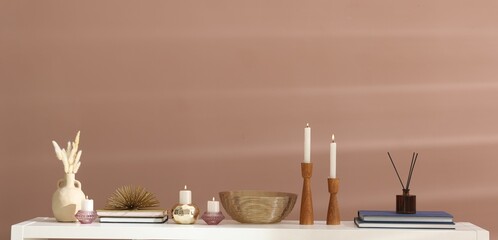 Candlesticks with burning candles and other decor on white wooden table near light brown wall indoors, space for text