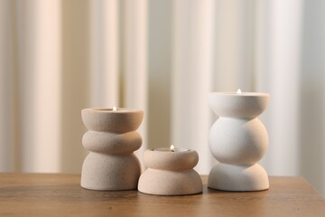 Stylish candlesticks with burning candles on wooden table against beige curtain indoors