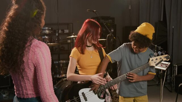 Two positive multiethnic teenagers chatting during band rehearsal, Afro American boy pulling strings of girls electric guitar, she hitting his hand in professional studio