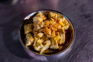 Close-up of Taiwanese fried stinky tofu with pickled cabbage and chili sauce in a round metal dish on a market table