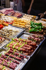A close-up of skewered raw meats and vegetables on trays under bright lights at a Taiwanese night market, showcasing a variety of barbecue options