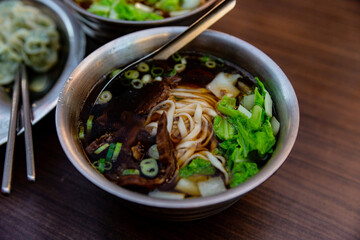 Taiwanese beef noodle soup served in a stainless steel bowl, with flat noodles, green vegetables, and slices of braised beef in rich broth