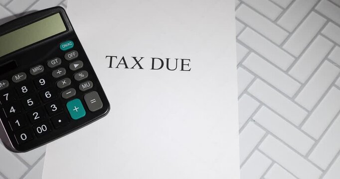 A black calculator rests on a white sheet of paper boldly labeled &ldquo;TAX DUE,&rdquo; set against a herringbone-patterned surface.