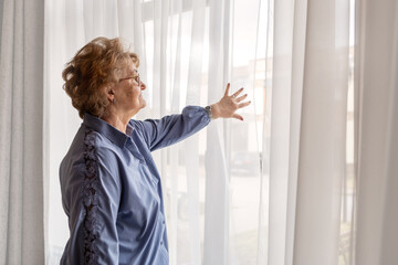 Senior woman in light blue clothes standing near window and gently looking outside through sheer curtains, thoughtful and peaceful moment in cozy home, symbolizing solitude, reflection, and calmness