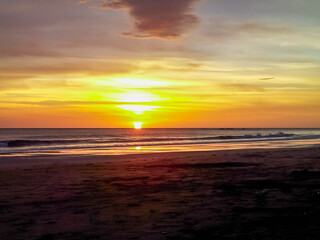 Fototapeta premium Brilliant sun dips below the ocean horizon, painting the sky with vibrant yellow and orange hues. Its reflection shimmers on the gentle waves washing onto dark sand beach along the Costa Rican coast
