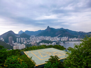 Obraz premium Prominent helipad offers a unique viewpoint overlooking Rio de Janeiro's stunning cityscape and Guanabara Bay, with iconic Sugarloaf and Corcovado mountains beneath moody sky in Brazil. Sweeping vista