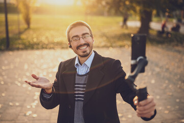 Fototapeta premium Male influencer explaining his business success through video blogging, smiling and gesturing while recording with his smart phone and gimbal in a park.