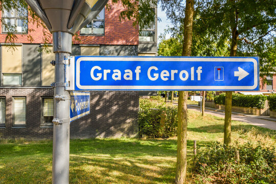 A blue directional sign indicating the location 'Graaf Gerolf' situated in an urban area with greenery and modern buildings in the background.