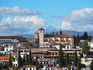 Traditional Albaic&iacute;n neighborhood in Granada, Spain, with whitewashed houses, church towers, and a mountainous backdrop under a partly cloudy sky.