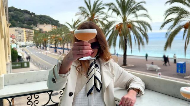 Stylish woman in sunglasses and a scarf holds a cocktail on a balcony with a scenic view of palm-lined Promenade des Anglais in Nice, France.
