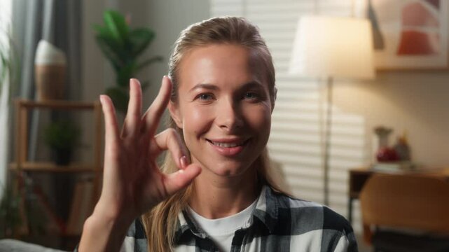 Caucasian woman adult lady homeowner looking at camera showing ok gesture okay sign blink eye blinking at home female in living room smiling demonstrate good result hand symbol fine loan mortgage