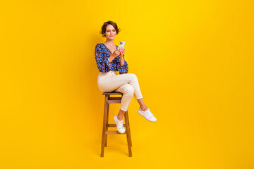 Full length photo of chill lady with smartphone texting her facebook friends sitting wooden bar chair isolated on yellow color background