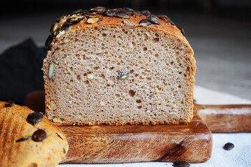 Loaf of bread with pumpkin seeds close-up on wooden board and cotton towel near knife on grey background