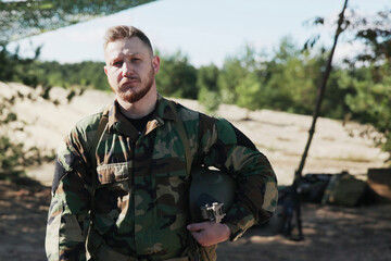 The weary soldier, clad in camouflage, stands with his helmet under his arm, looking directly into...