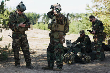 A group of soldiers prepares for a field patrol, setting up strategy and location, following a map on a computer and tablet, while the troop leader secures his helmet.