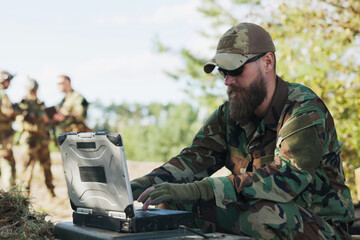 A military unit, equipped for combat, waits in the distance for the commander, who sits at the computer, entering mission details and sending crucial data to the base.