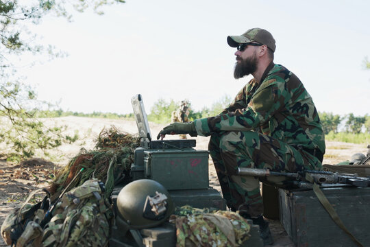 In the field, the army commander works on a computer, preparing reports for the base. A soldier arrives with a map, and they discuss strategy as others guard the camp in the background.