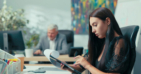 A young woman, company office worker, assistant to an elderly boss, sits at desk analyzing reports...