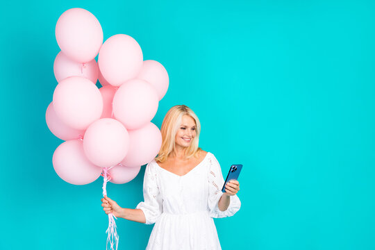 Blonde woman holding pink balloons on a turquoise background while inspecting her phone, exuding happiness and charm