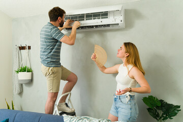 Young couple fixing an air conditioning unit while woman holds hand fan