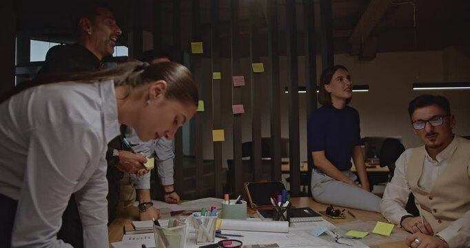 Diverse business team collaborates on a project, using sticky notes and blueprints in a modern office. They share ideas and give high fives.