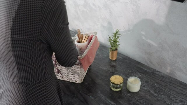 Partial side view of housewife in checkered dress unpacking basket and placing items like glass jar and candle on marble kitchen table near small potted plant under soft daylight in cozy modern home