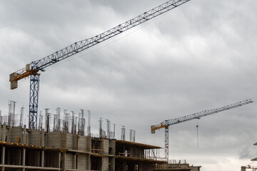 Modern construction site with two tower cranes and unfinished concrete building under dramatic cloudy sky. Image ideal for illustrating urban development, architecture, and infrastructure projects. © Mike Loik