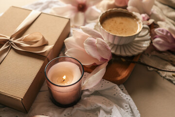 A cozy still life featuring a cup of coffee on a wooden tray, a burning candle in a glass holder, a kraft gift box with a satin ribbon, and magnolia blossoms on soft fabrics