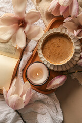 Lit candle in a glass jar with soft magnolia flowers, a gift box, and a coffee cup in warm pastel light on white lace fabric