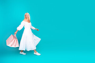 Graceful woman in white summer dress carrying shopping bags stands on vibrant teal background...