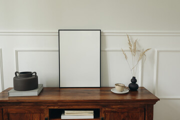 Scandinavian interior. Modern living room design. Blank picture mock up poster frame. Mahogany wooden dresser sideboard. Black round vases with dry grass. Cup of tea, coffee. White wall background.